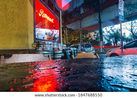 Restaurant facade, bright red sign with beer barrels on the sidewalk in a rainstorm. Wet corner bar table.