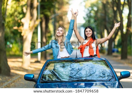 Two young women have fun with raised hands in convertible car on summer street