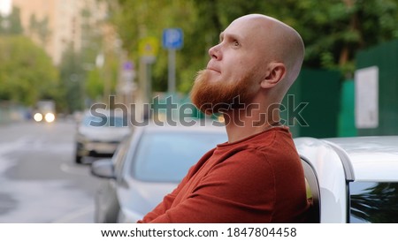 A young brutal bald guy with a well-groomed red beard and a sweater stands in the street, leaning against a car in a pensive state. Reflections on life. Summer evening. Royalty-Free Stock Photo #1847804458