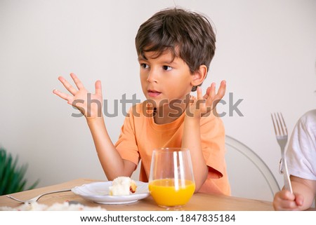 Portrait of cute little boy eating birthday cake and drinking orange juice. Adorable child sitting at table in dining room, rising hands and looking away. Childhood, celebration and holiday concept