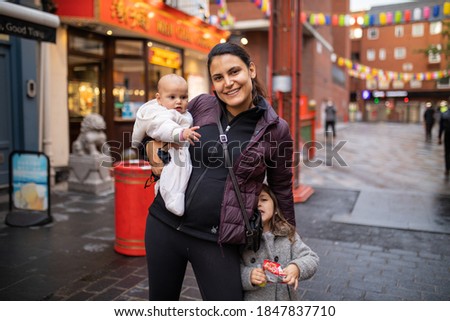 Picture of a smiling woman standing alongside her little daughter and carrying her baby with her right arm, with an alley from Chinatown as background