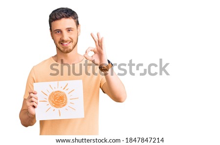 Handsome young man with bear holding sun draw doing ok sign with fingers, smiling friendly gesturing excellent symbol 