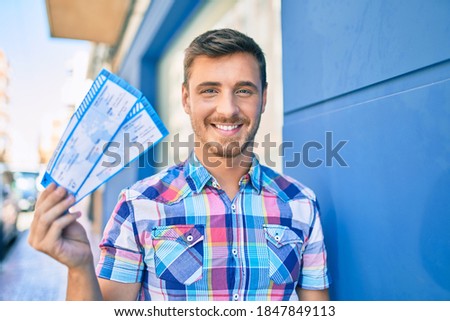 Young caucasian man smiling happy holding boarding pass ticket leaning on the wall at the city.