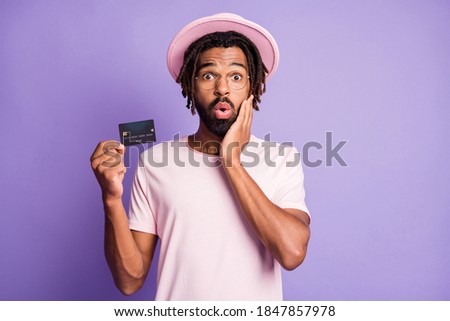 Photo portrait of shocked man holding credit card in one hand touching face cheek isolated on vivid purple colored background