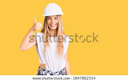 Beautiful caucasian woman with blonde hair wearing hardhat and painter clothes smiling happy and positive, thumb up doing excellent and approval sign 