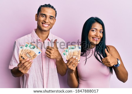 Young latin couple holding 50 euro banknotes smiling happy and positive, thumb up doing excellent and approval sign 