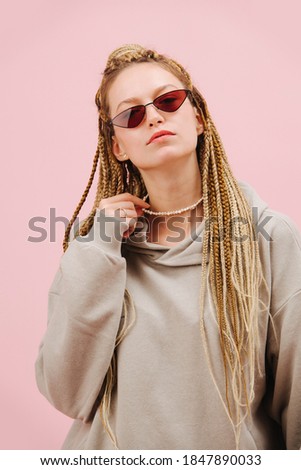 Cool looking young woman in narrow sunglasses with stylish blond afro braids over pink background. Looking down with chin up, arrogant expression. Holding pearl necklace.