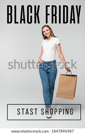 joyful young woman holding shopping bag while standing with hand in pocket near black friday lettering on grey