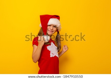 Kid wearing Santa hat holding her fists clenched in victory and joy, cheering and looking towards Christmas presents, isolated on yellow colored background