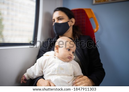Picture of a woman wearing a black face mask sitting next to a window in a bus and holding her baby daughter who looks absently at her left side