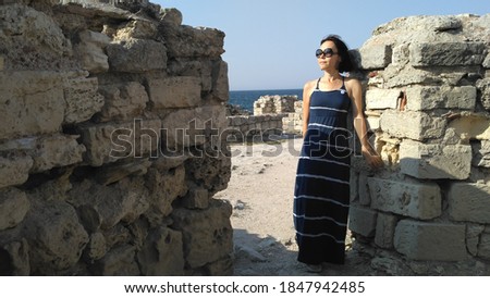 Lonely female in a long blue dress on the background of the Mediterranean landscape. A beautiful young woman in sunglasses travels and enjoys and admiring the ancient city ruins and historical place.