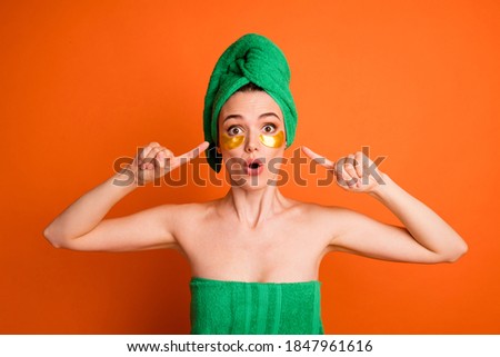 Photo portrait of surprised woman pointing two fingers at golden eye patches wearing green towel isolated on vivid orange colored background