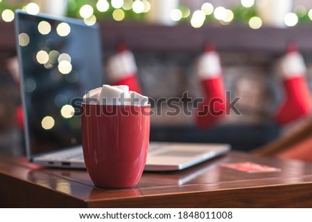 Credit card, laptop on the table for making order. Cup of hot cocoa and marshmallow at christmas fireplace with decoration of light bulbs. Close up.