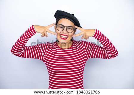Young beautiful brunette woman wearing french beret and glasses over white background Doing peace symbol with fingers over face, smiling cheerful showing victory