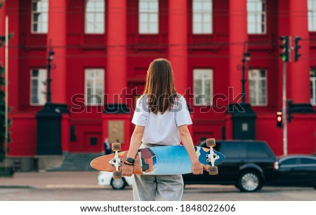 Back of a stylish skater girl with a longboard behind her back stands on a background of red architecture. Background