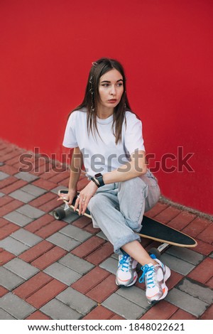 Stylish woman in street clothes sits on a longboard near a red wall and poses for the camera looking to the side.