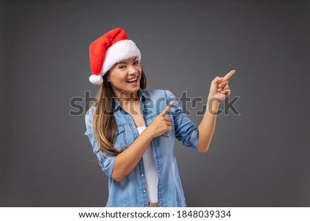 Young smiling woman with santa's hat dressed casual standing in front of gray background and pointing up with fingers.