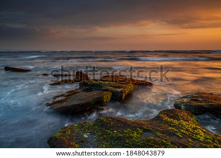 Beautiful seascape. Beach with stones covered by seaweeds. Low tide. Composition of nature. Motion water. Cloudy sky with sunlight. Slow shutter speed. Soft focus. Mengening beach, Bali, Indonesia
