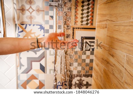 Man's hand checking the water temperature in the shower using a hand-held shower wall in a bathroom with beautiful modern ceramic tiles.