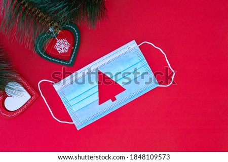 Medical protective mask with cut-out silhouette of Christmas tree on red background.With Christmas tree branches near the mask. Christmas and Happy New Year concept, coronavirus concept.
