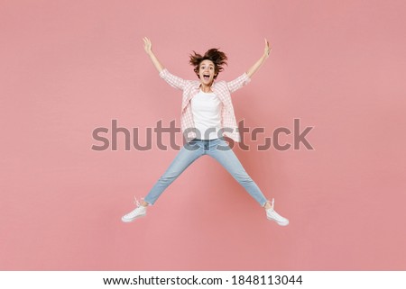 Full length of excited cheerful young brunette woman 20s wearing casual checkered shirt jumping spreading hands and legs keeping mouth open isolated on pastel pink colour background, studio portrait