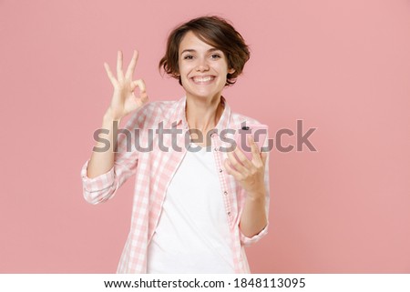 Smiling young brunette woman wearing casual basic checkered shirt standing using mobile cell phone typing sms message showing OK gesture isolated on pastel pink colour background, studio portrait