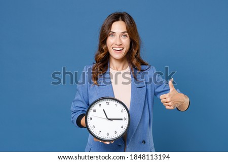 Smiling cheerful funny young brunette woman 20s wearing basic casual jacket standing holding in hands clock showing thumb up isolated on bright blue colour wall background studio portrait