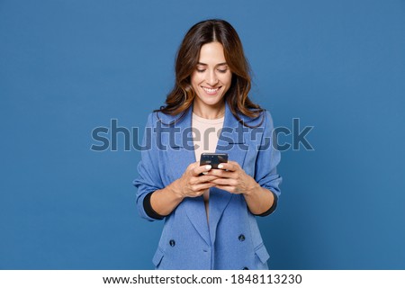 Smiling funny beautiful attractive young brunette woman 20s wearing basic jacket standing using mobile cell phone typing sms message isolated on bright blue colour wall background, studio portrait