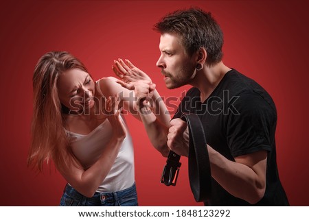 A man hits a defending woman with a belt during a scandal. Domestic violence against a girl. The husband mocks his wife. Quarrel in the family. Abusive relationships. Royalty-Free Stock Photo #1848123292