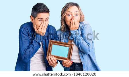 Couple of women holding empty frame laughing and embarrassed giggle covering mouth with hands, gossip and scandal concept 