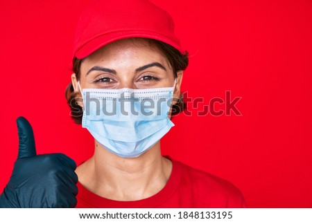 Young hispanic girl wearing delivery uniform and covid-19 safety mask smiling happy and positive, thumb up doing excellent and approval sign 