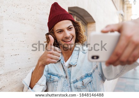 Photo of a happy young curly man walking outdoors by street and taking a selfie by by mobile phone with thumbs up gesture
