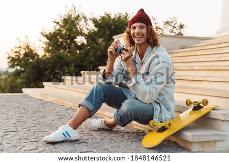 Picture of positive happy young curly man scater sitting outdoors and holding camera