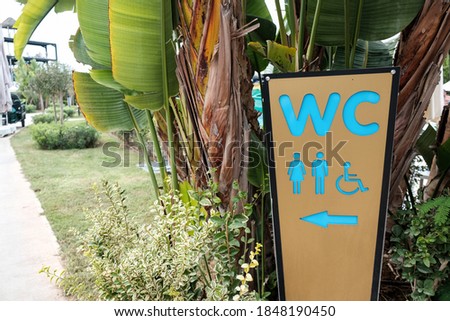 WC sign to a toilet room or washroom or bathroom or restroom for men and women and invalids or disabled people. Beautiful green tropical trees and bushes during summer. Board with a direction arrow.