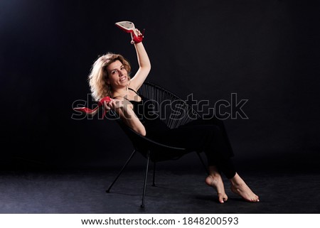 Young blond curly woman, sitting on chair in front of black background. Full-length portrait of female, wearing black clothes, holding red high-heeled shoes. Love yourself concept. Studio photography.