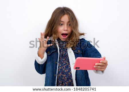 Wow!! excited Little caucasian girl with beautiful blue eyes wearing denim jacket standing over isolated white background showing mobile phone with open hand gesture 