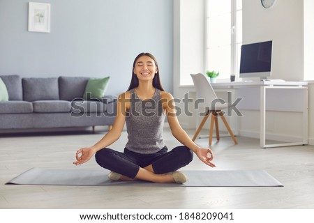 Happy smiling young woman practicing meditation in easy pose taking break from work in home office. Positive Asian girl sitting legs crossed on yoga mat reaching zen, enjoying stress relief exercise Royalty-Free Stock Photo #1848209041