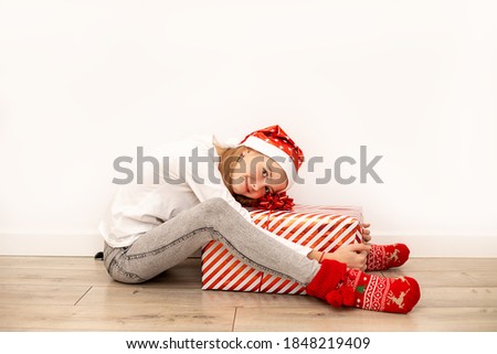 A beautiful girl in casual clothes with Christmas accessories sits on the floor and hugs a large gift box with a red ribbon and a bow. Full-length photo. Copy space