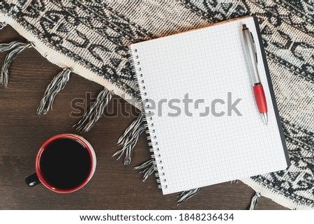 Blank notebook with a pen, a glass of coffee and a woolen warm tablecloth on a wooden table. View from above.