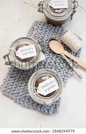 Three chocolate mousse covered jars on a knitted blue cloth next with a couple of spoons in the middle and three tags on the jars..