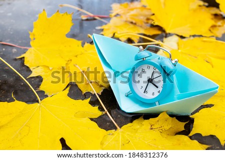 blue alarm clock floating on a paper ship over water surface with autumn leaves. wasting time concept. time to travel conceptual.