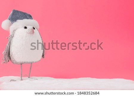 A closeup of a small penguin toy with a Christmas hat on artificial snow against a pink background
