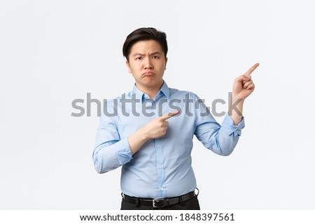 Business, finance and people concept. Disappointed gloomy asian male office worker, salesman in blue shirt complaining something bad, showing way, pointing fingers upper right corner