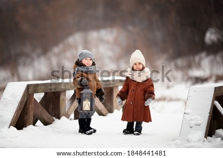 A boy and a girl stand on a snow-covered wooden bridge. The boy holds a lantern in his hands. Children in warm sheepskin coats and hats. Image with selective focus.
