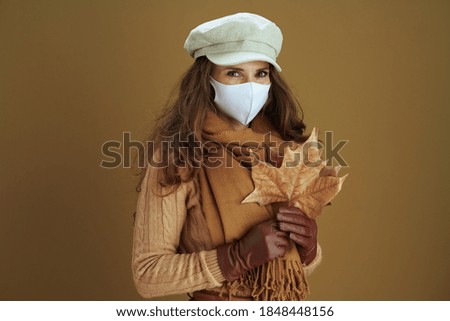 Life during coronavirus pandemic. Portrait of 40 years old housewife in scarf with white medical mask and yellow autumn maple leaves isolated on beige.