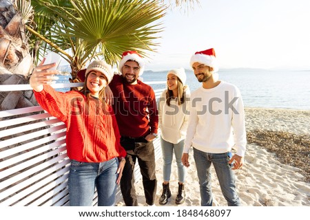 Group of young caucasian friends with Santa Claus hat having fun taking a selfie at the beach in winter for the Christmas holidays - Two couple of people using technology to sharing their journey