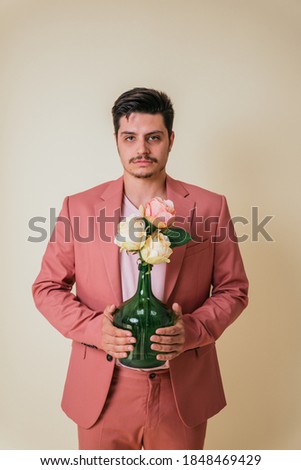 Handsome young man looking at the camera in a photo studio on a yellow background holding a bottle with flowers dressed in a pink suit and T-shirt.