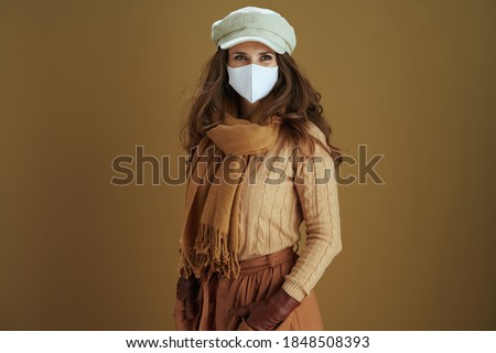 Hello autumn. pensive modern middle aged housewife in scarf with white medical mask looking into the distance isolated on brown background.