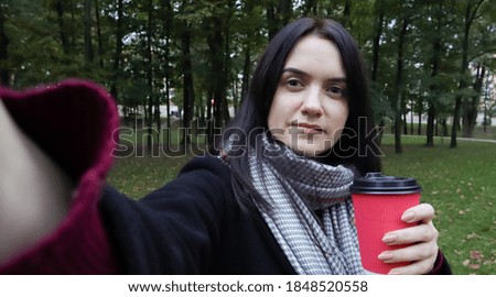 Beautiful young caucasian woman in a coat with a paper cup of takeaway coffee taking a selfie or taking pictures of herself for a blog with a smartphone outdoors in an autumn park.
