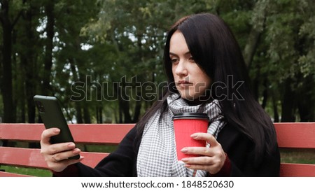 Beautiful young caucasian woman in a coat with a paper cup of takeaway coffee taking a selfie or taking pictures of herself for a blog with a smartphone outdoors in a park sitting on a bench.